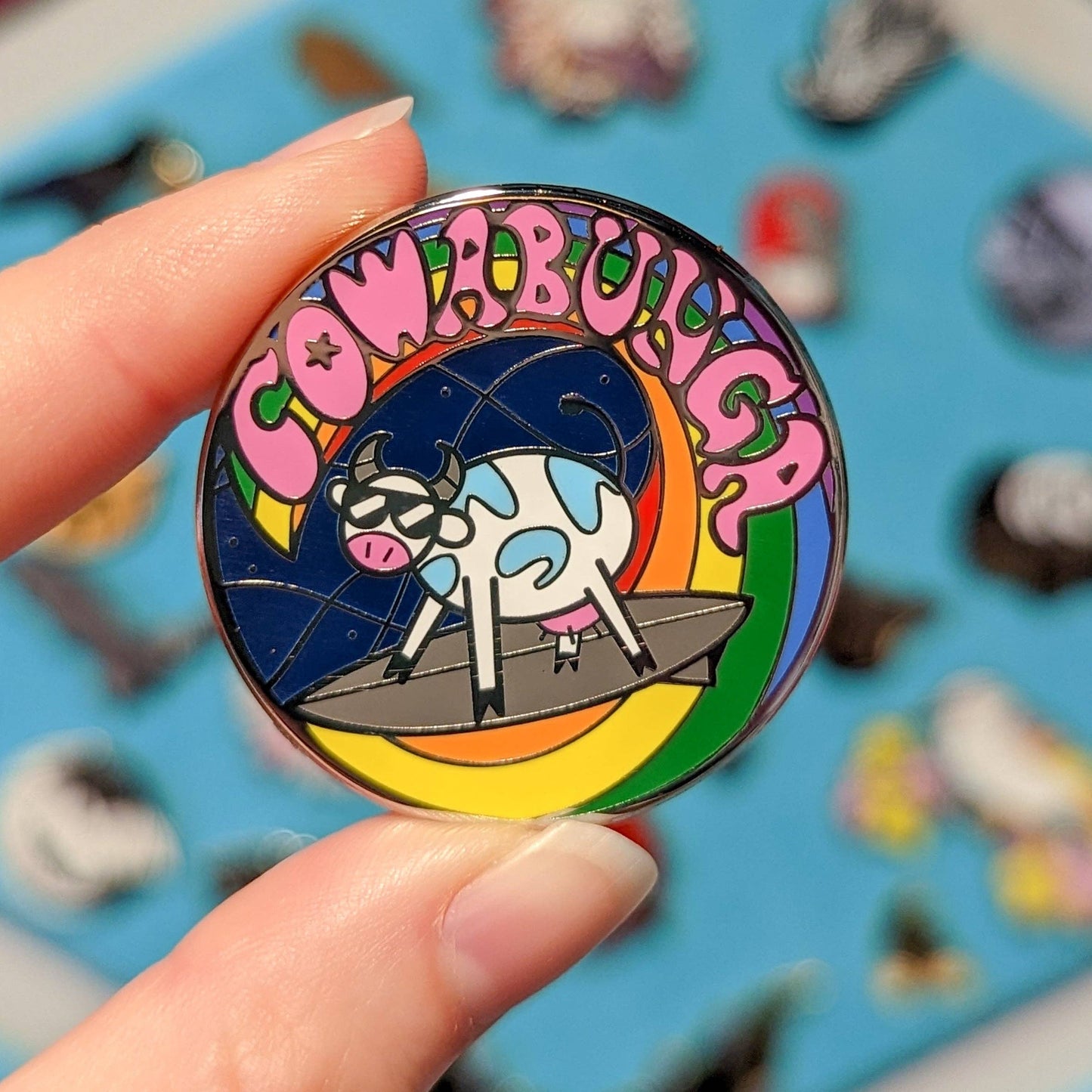 a hand holding a Cowabunga Pride enamel pin with sunglasses-wearing cow surfing on a rainbow wave