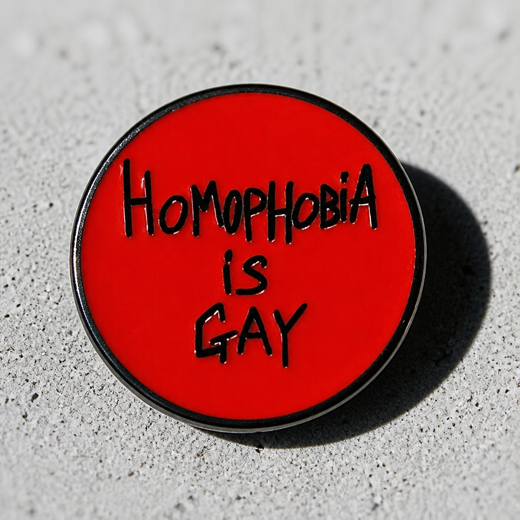 Round red hard enamel pin featuring the phrase "Homophobia Is Gay" in black hand-lettered text, displayed on a concrete surface
