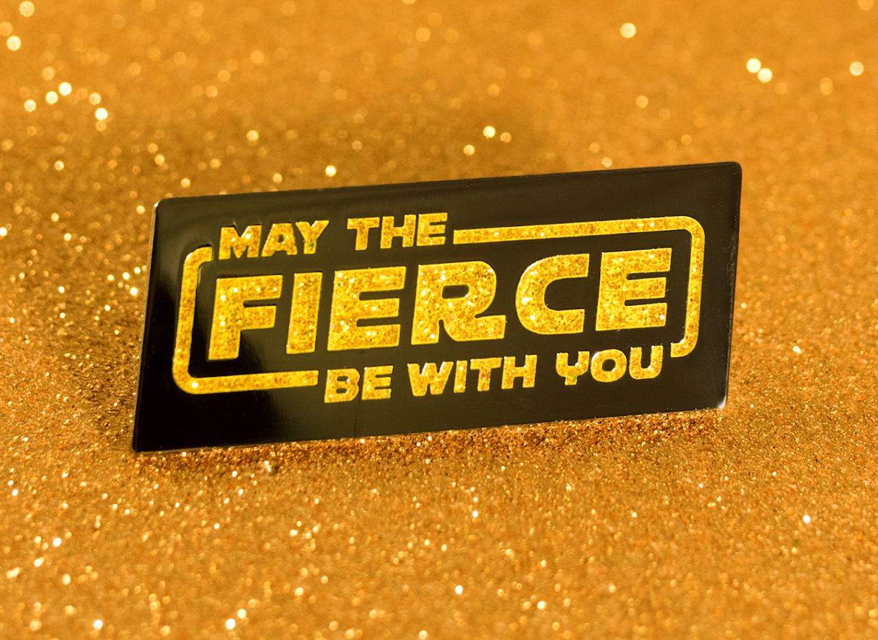 Black enamel pin with glitter gold lettering reading May the Fierce Be With You