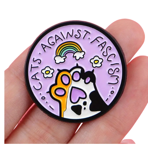 Pastel enamel pin reading “Cats Against Fascism” with rainbow and cat paw artwork held in a hand