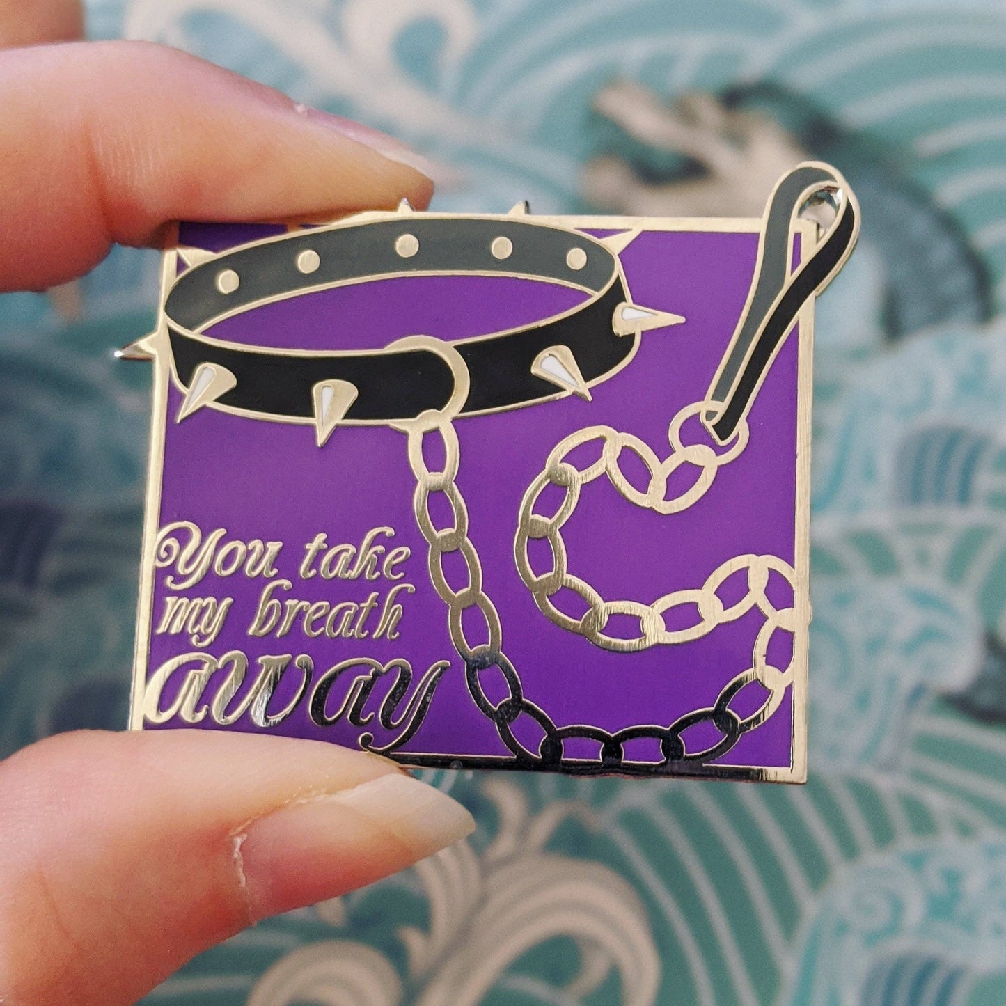 a hand holding a You Take My Breath Away enamel pin with black spiked choker and chain on purple background