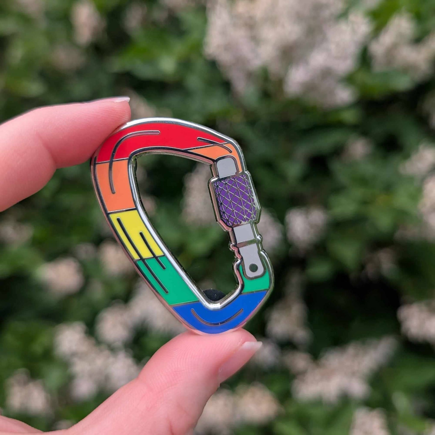 hand holding a Rainbow Pride enamel pin shaped like a colorful silver-tone carabiner