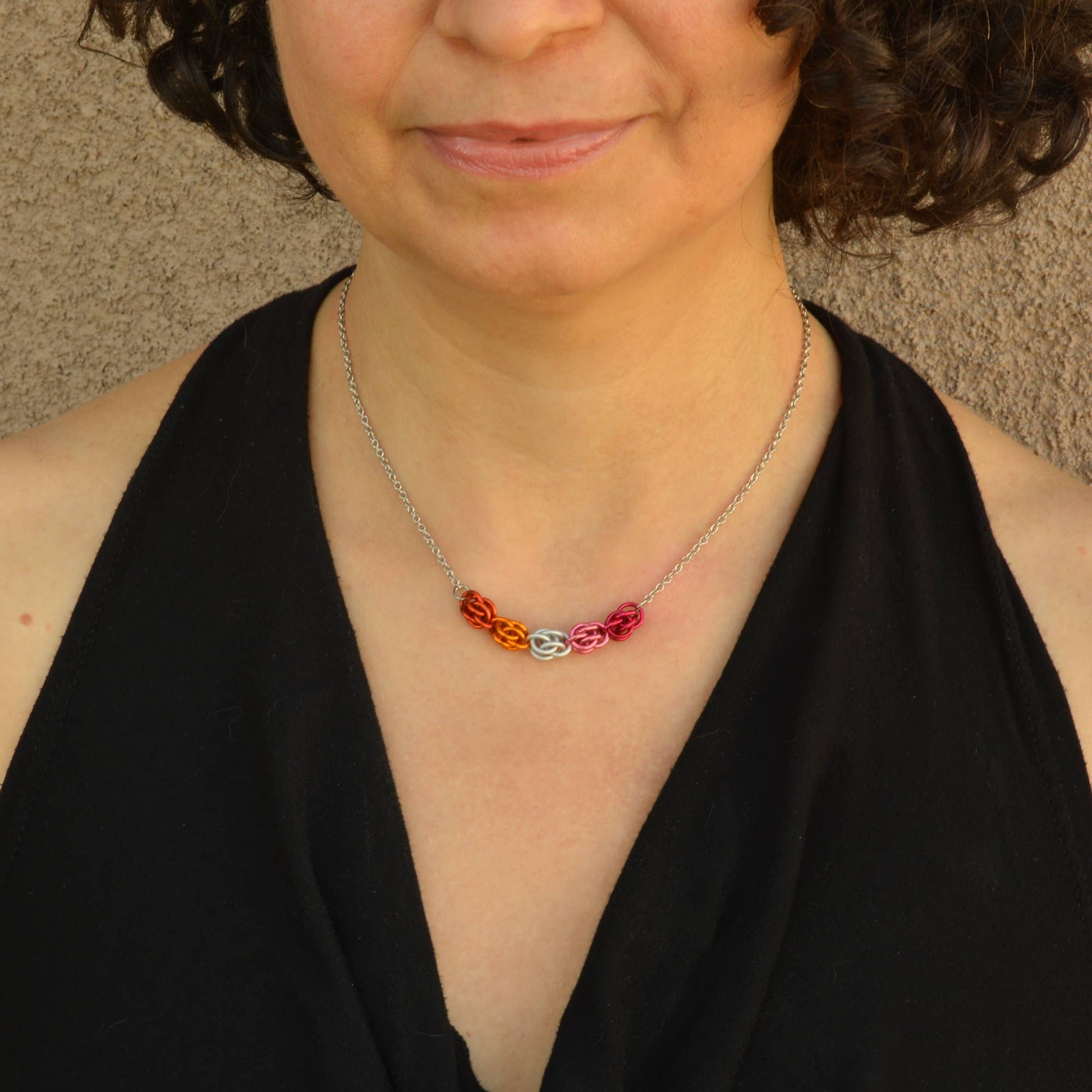 a close-up of a woman wearing a Hand-linked lesbian pride chainmail necklace featuring orange, white, pink, and magenta aluminum rings on an adjustable stainless steel chain.