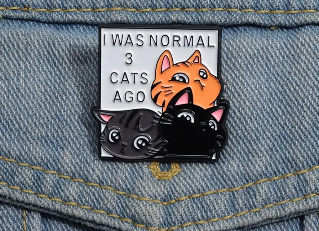 Enamel pin with cartoon cats and text 'I was normal 3 cats ago' on denim fabric
