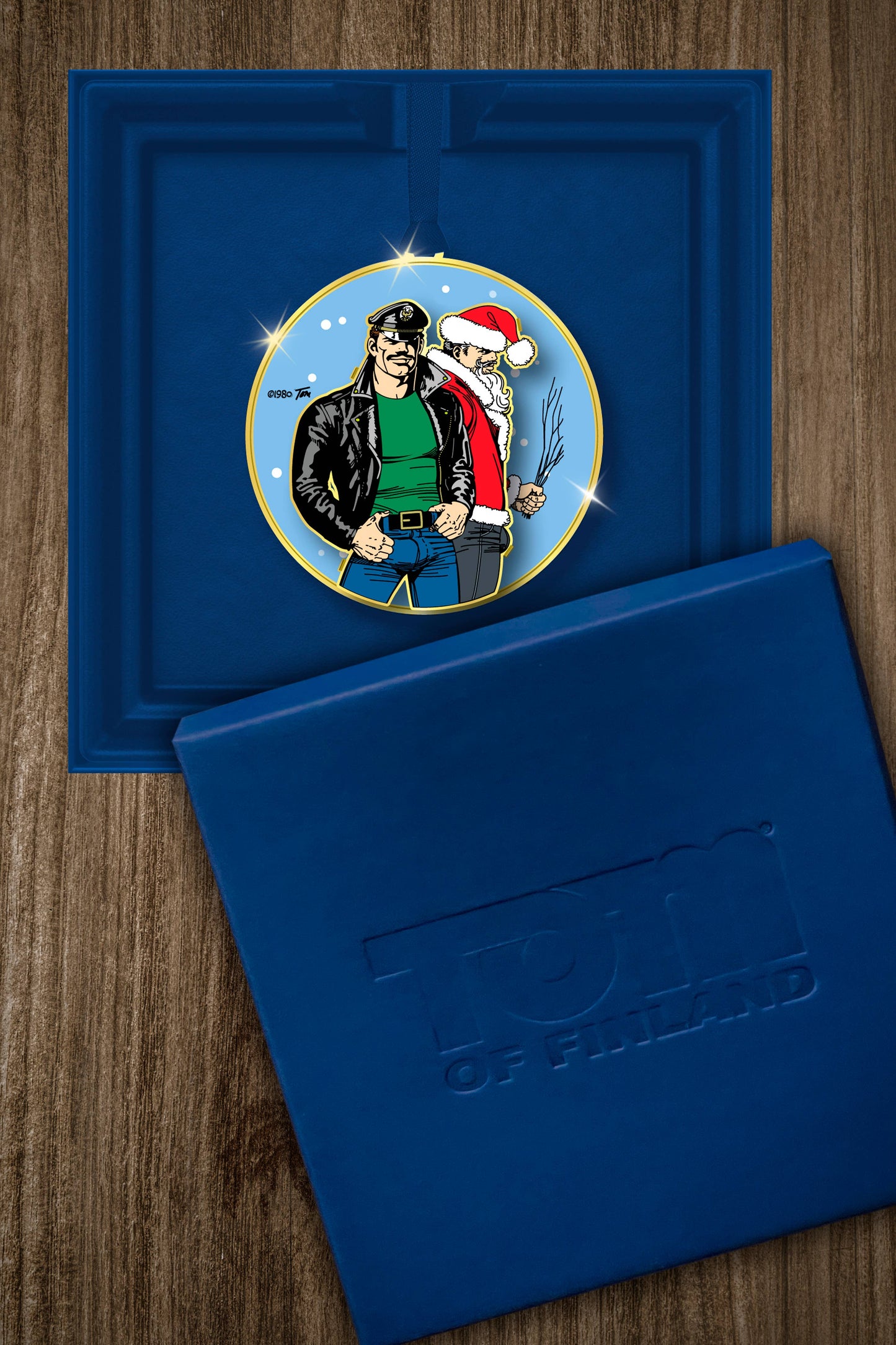 Limited edition Tom of Finland Leather & Santa metal ornament featuring a leather-clad character standing with Santa, presented in an embossed navy gift box.