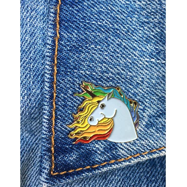 White unicorn enamel pin with flowing rainbow mane and gold-tone outline displayed on a blue denim pocket