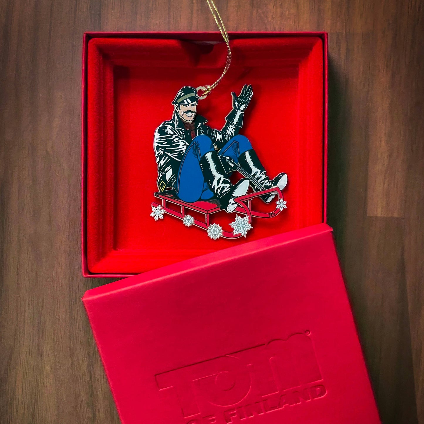 Limited edition Tom of Finland leather sled metal ornament featuring a leather-clad character riding a red sled, displayed in an embossed red gift box.