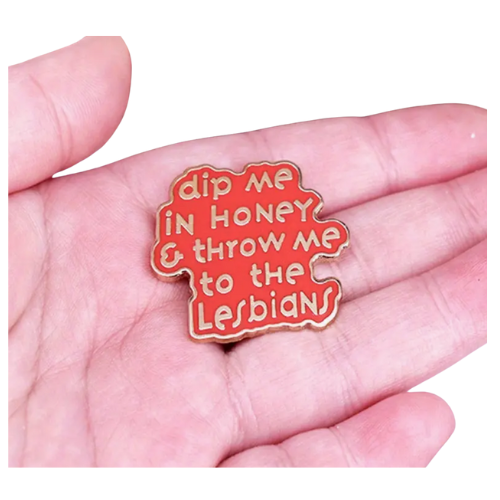 Pink enamel pin with the phrase “Dip Me in Honey & Throw Me to the Lesbians” held in a hand