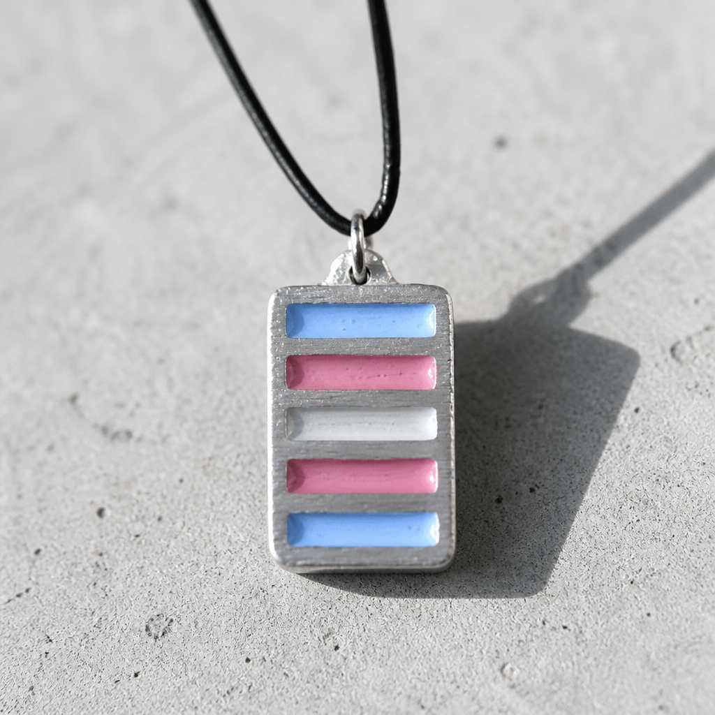 Handcrafted pewter pendant featuring the Transgender Pride flag in a contemporary geometric design on an adjustable black waxed cord on a cement surface