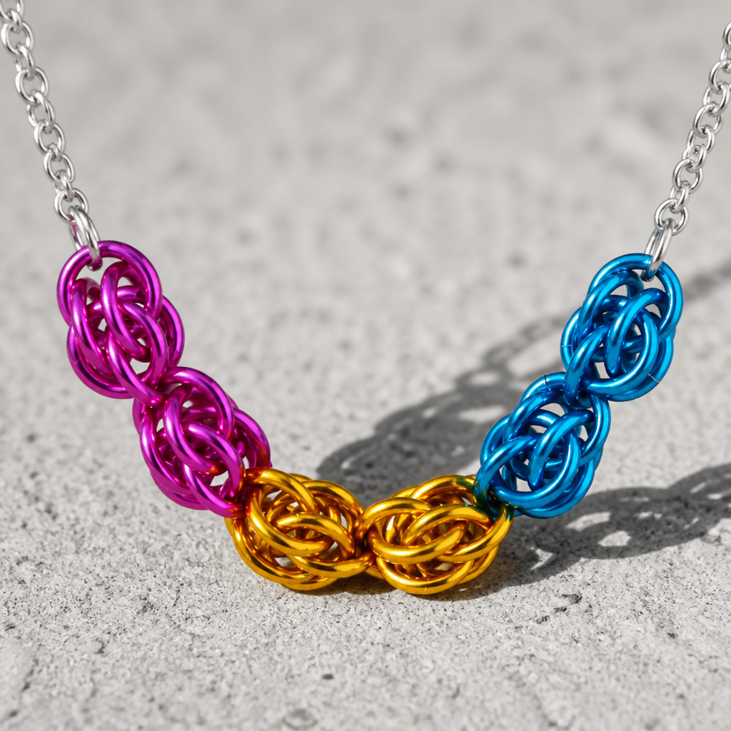 Hand-linked pansexual pride chainmail necklace featuring pink, yellow, and blue aluminum rings on an adjustable stainless steel chain on a concrete surface.
