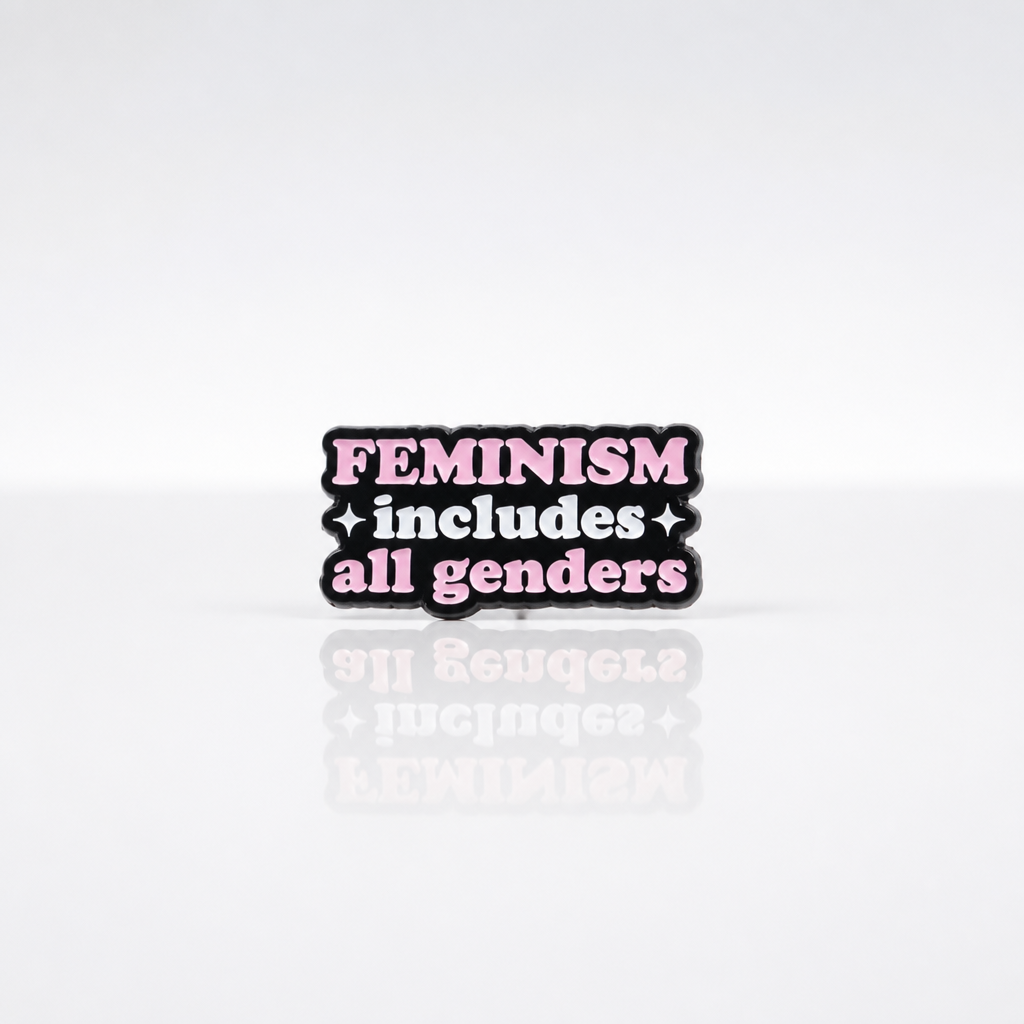 Pink and black enamel pin reading “Feminism Includes All Genders”