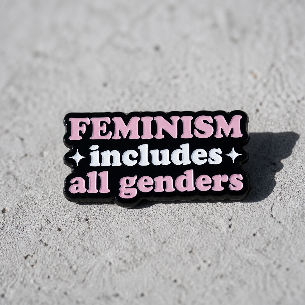 Pink and black enamel pin reading “Feminism Includes All Genders” on a concrete surface
