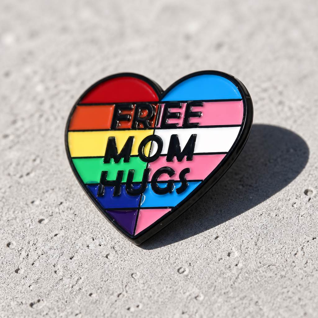 Heart-shaped Free Mom Hugs enamel pin with rainbow and trans pride flag colors on a concrete surface