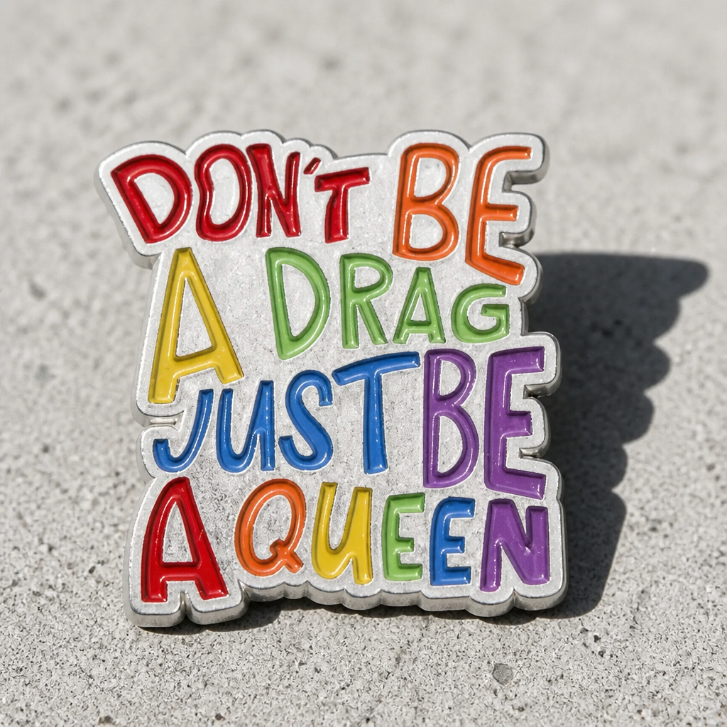 Colorful Don’t Be A Drag Just Be A Queen enamel pin with rainbow lettering on a concrete surface