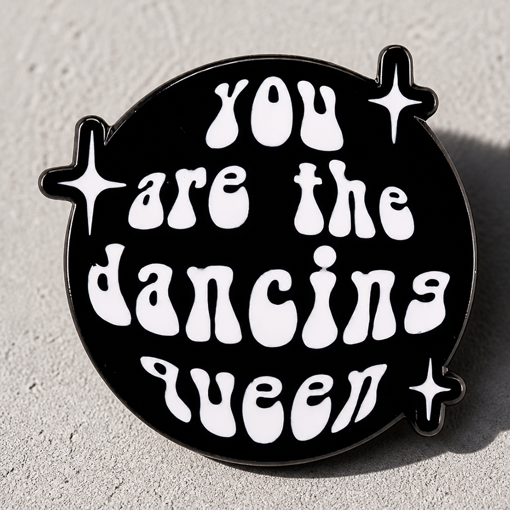 Retro Dancing Queen enamel pin with bold disco-inspired lettering and sparkles on a concrete surface