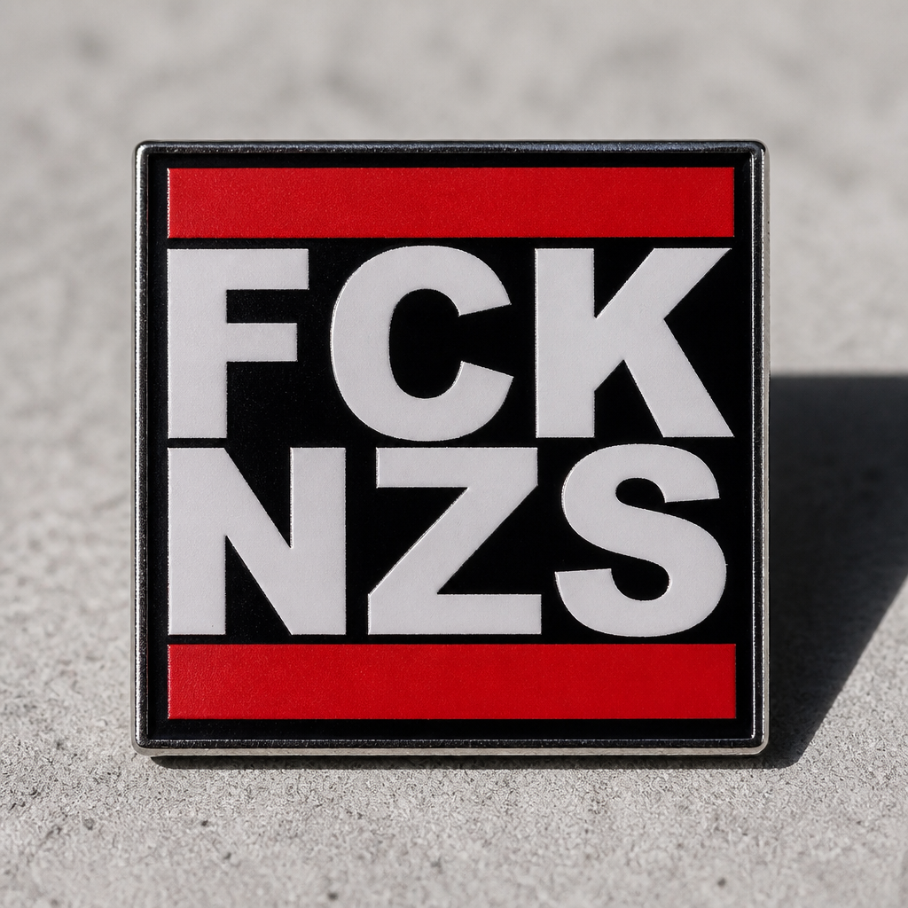 Black red and white enamel pin reading “FCK NZS” on a concrete surface