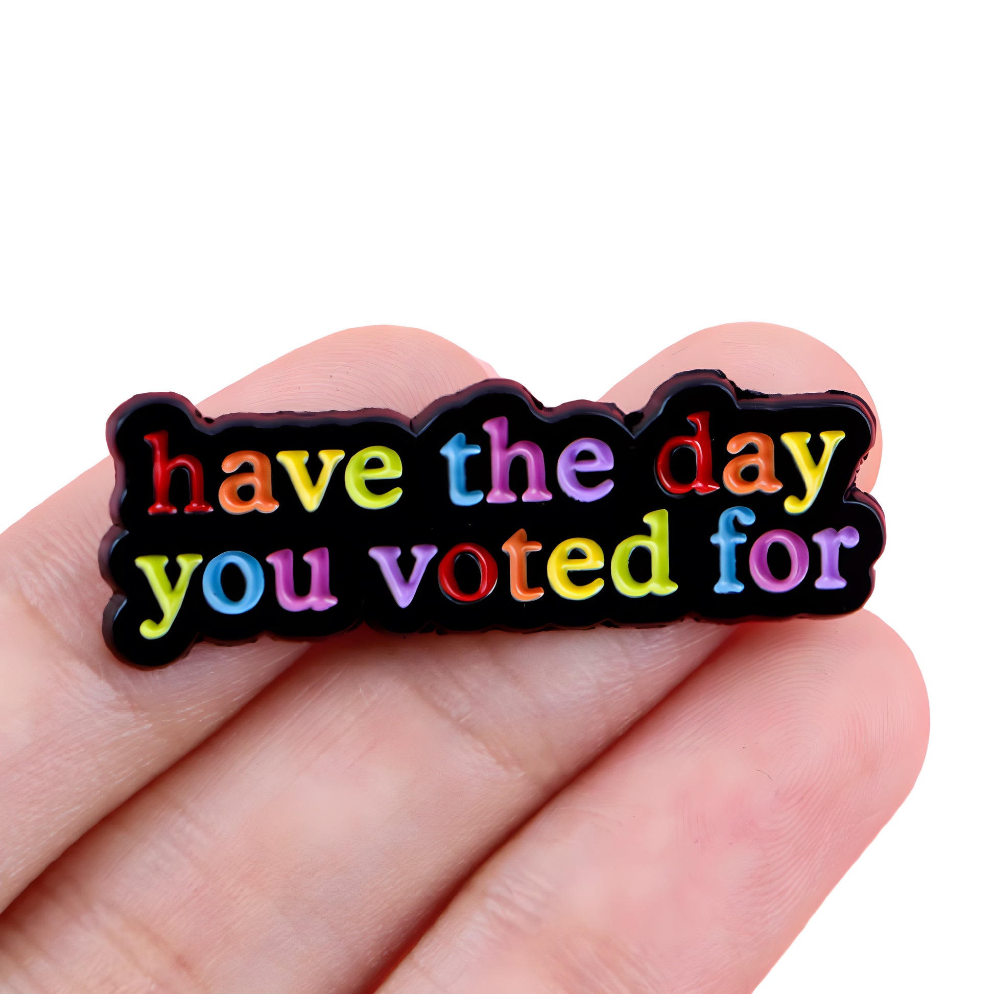 Rainbow enamel pin with “Have The Day You Voted For” text on black background held in a hand