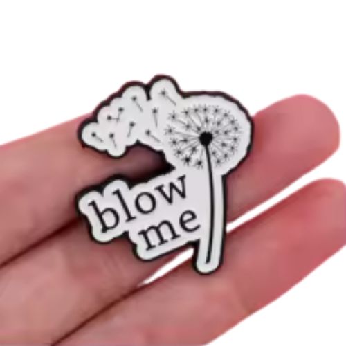 Black-and-white dandelion enamel pin with “Blow Me” text held in a hand