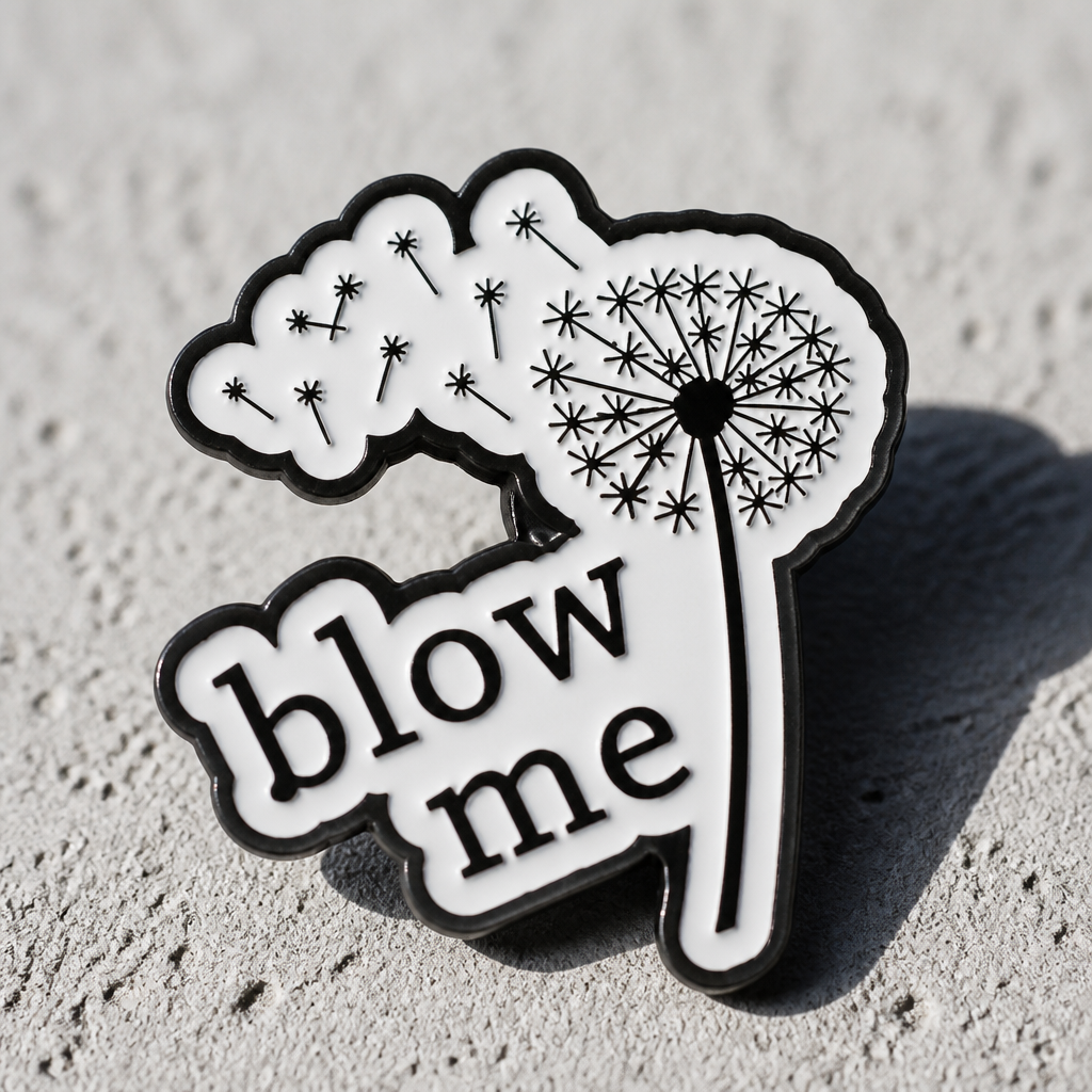 Black-and-white dandelion enamel pin with “Blow Me” text on a concrete surface