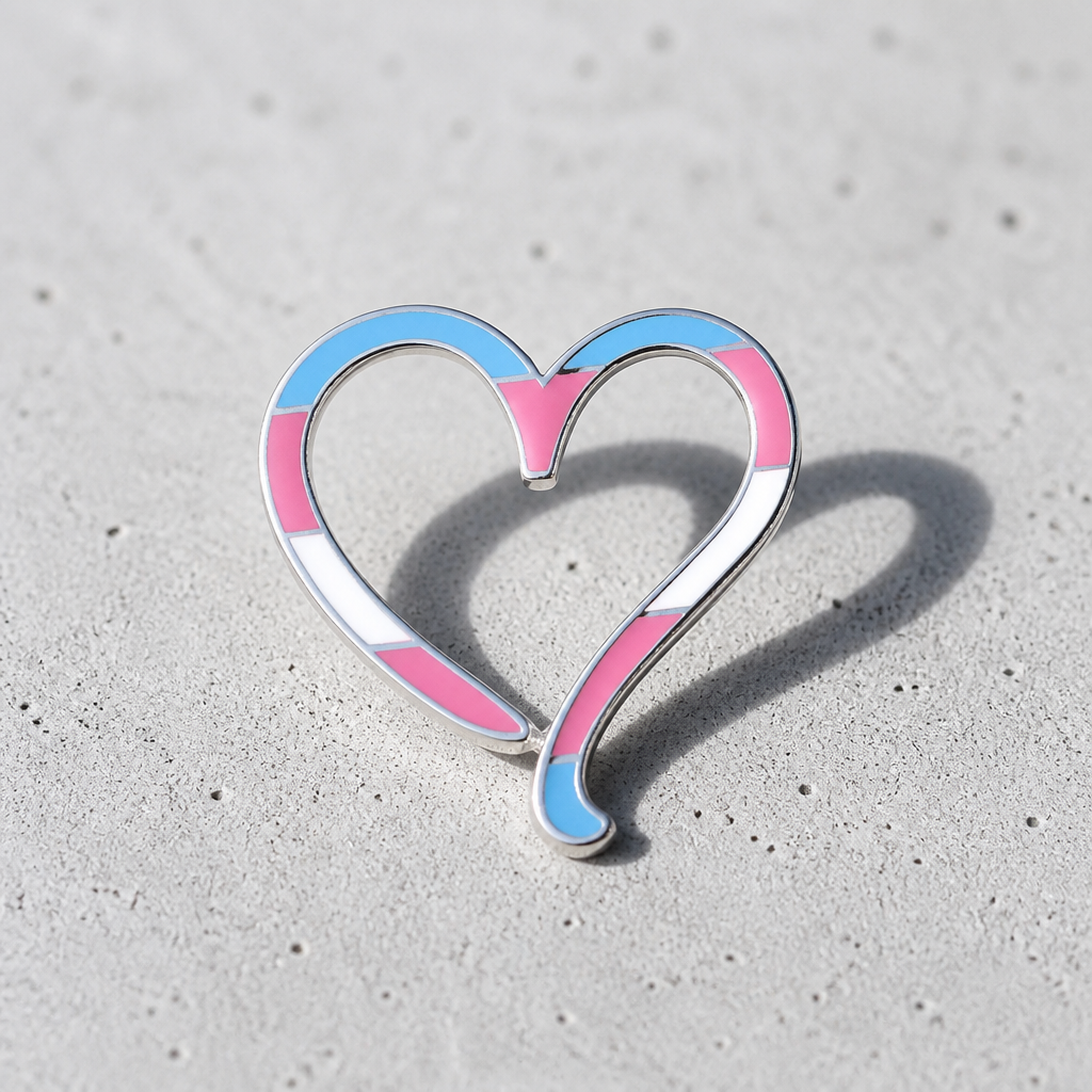 Hollow heart enamel pin with transgender pride flag colors and silver outline on a concrete surface