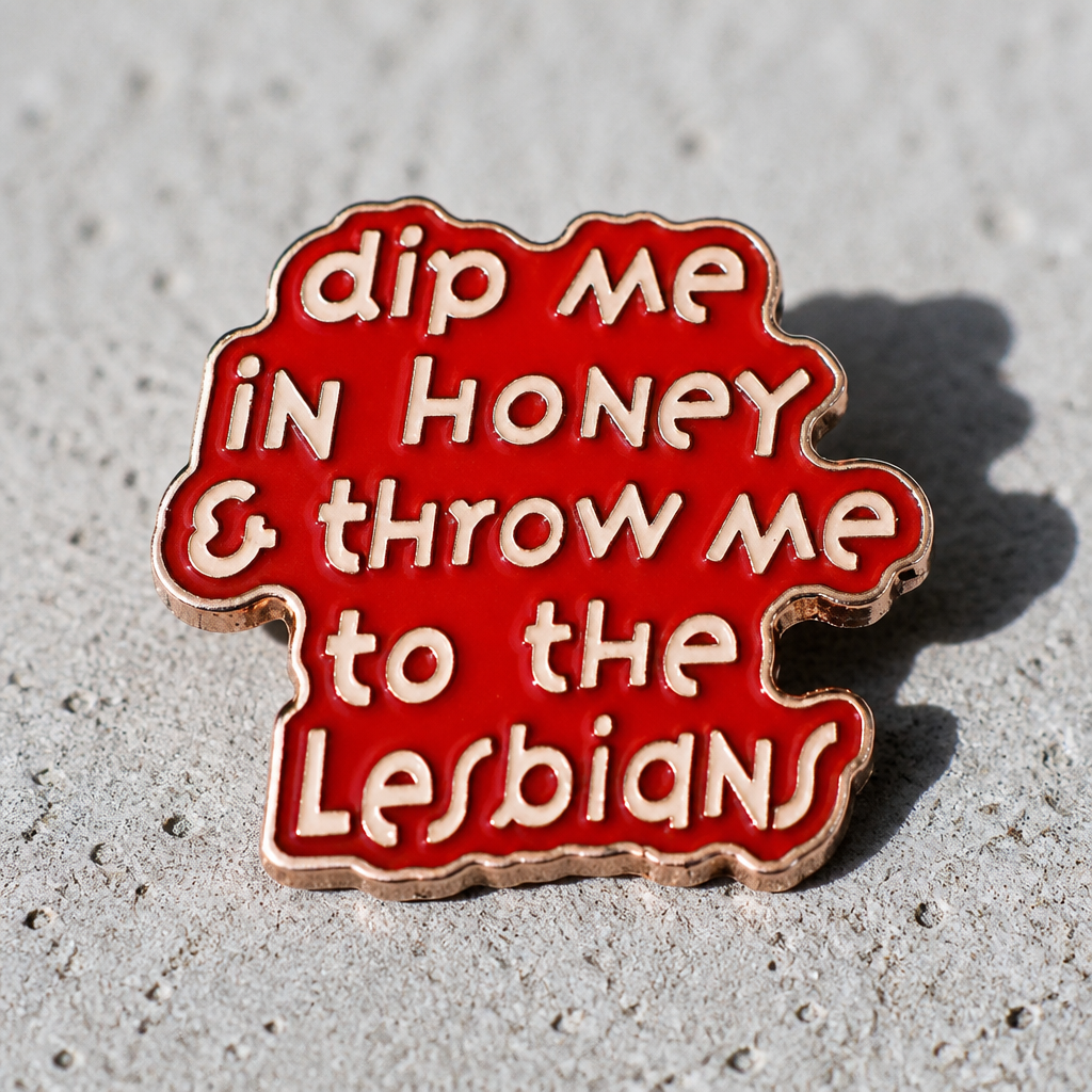 Pink enamel pin with the phrase “Dip Me in Honey & Throw Me to the Lesbians” on a concrete surface