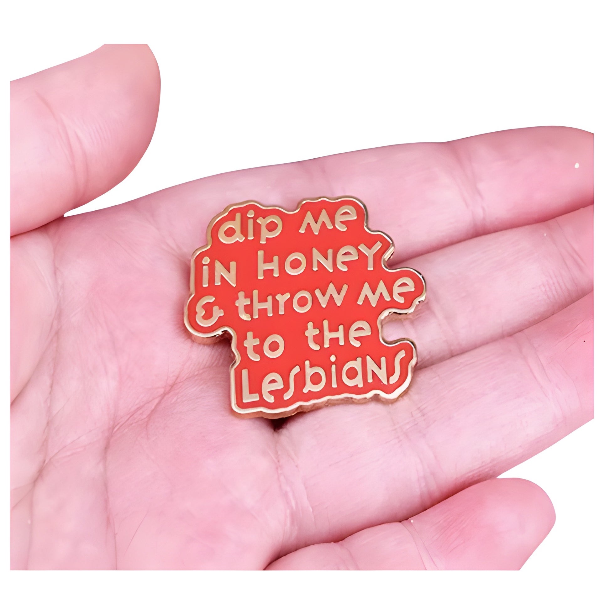 Pink enamel pin with the phrase “Dip Me in Honey & Throw Me to the Lesbians” held in a hand
