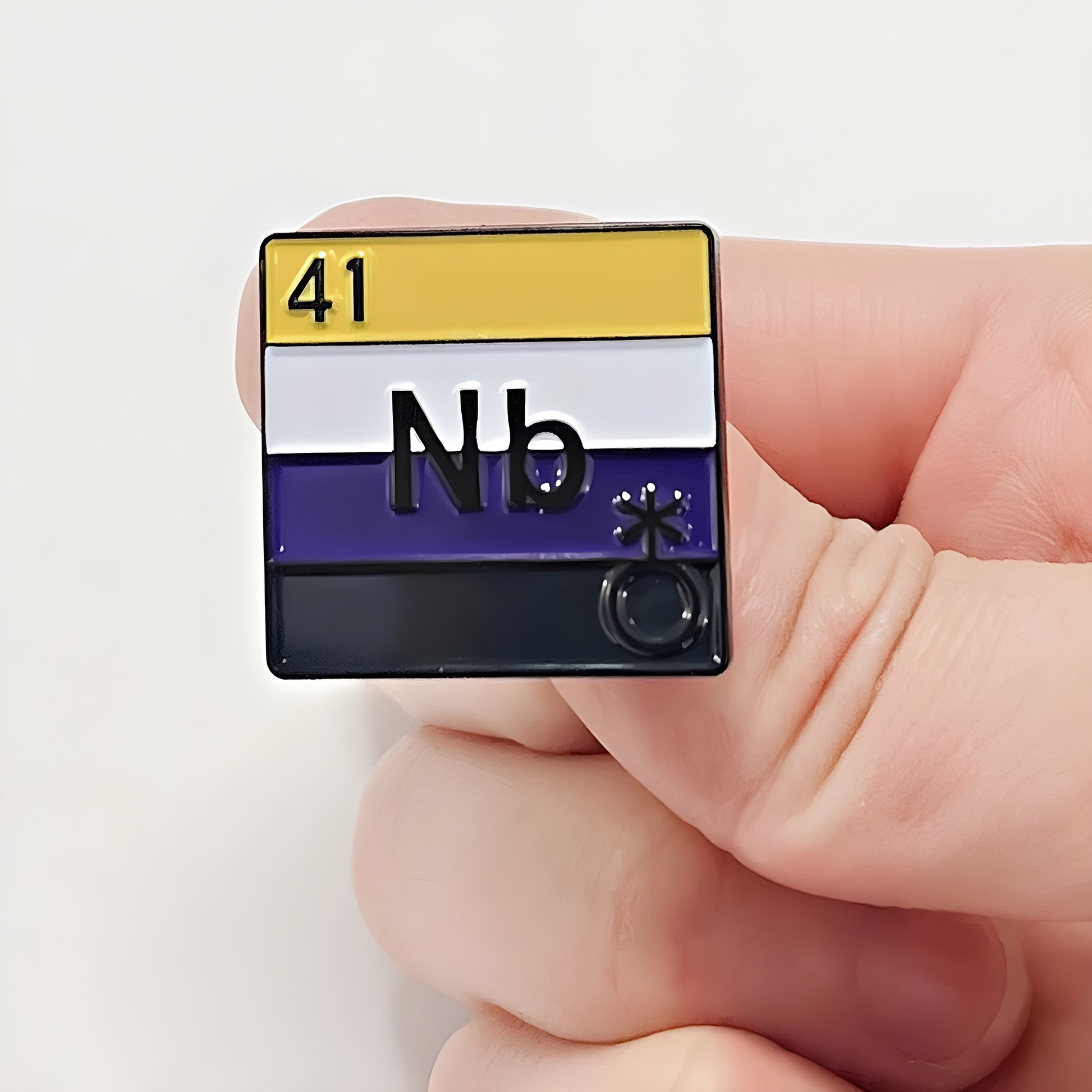 Hard enamel pin designed like a periodic table element featuring the Nonbinary Pride flag colors and the abbreviation "Nb." Held up between two fingers