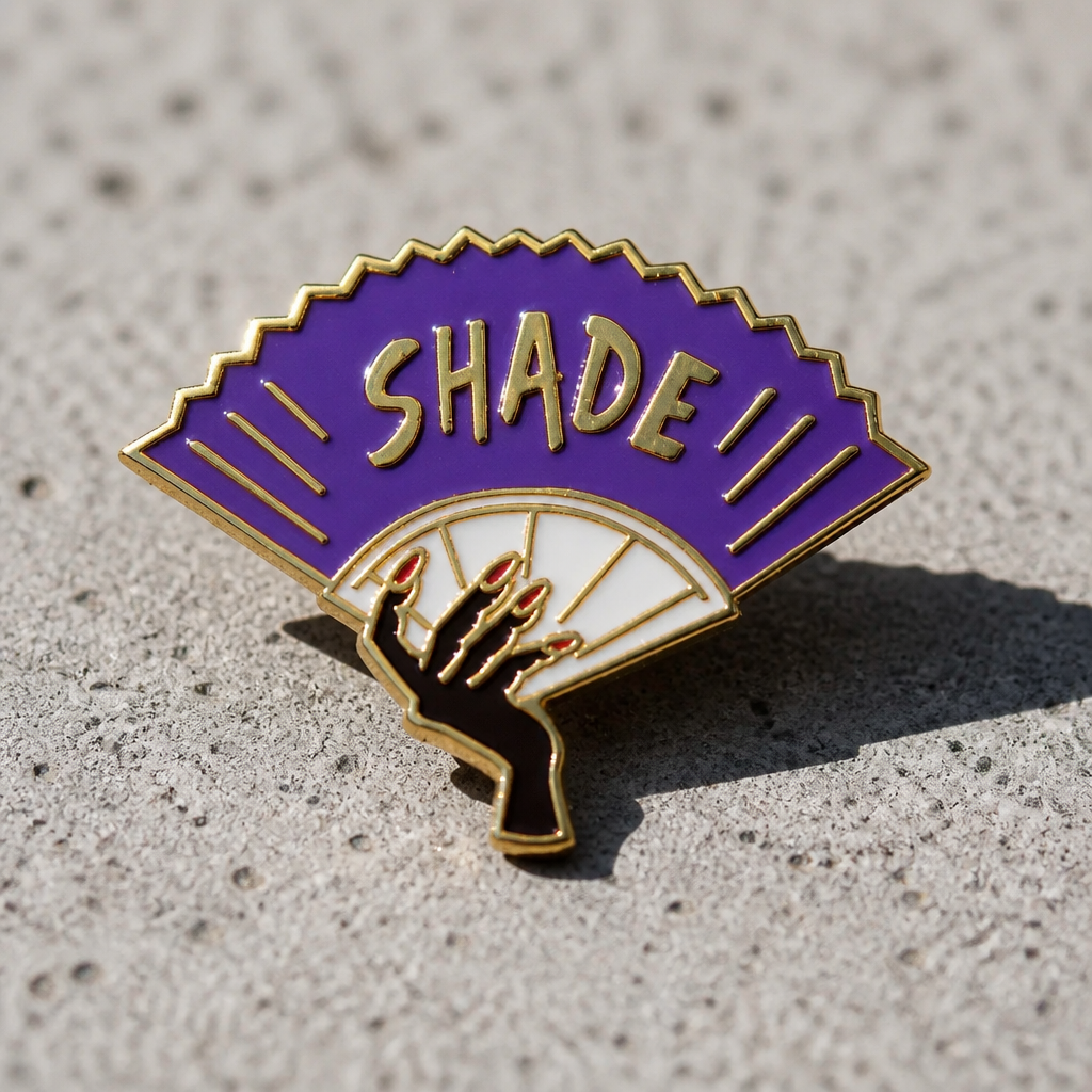 Purple Shade enamel pin featuring a dramatic folding fan design with gold-tone details on a concrete surface