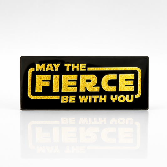 Black enamel pin with glitter gold lettering reading May the Fierce Be With You
