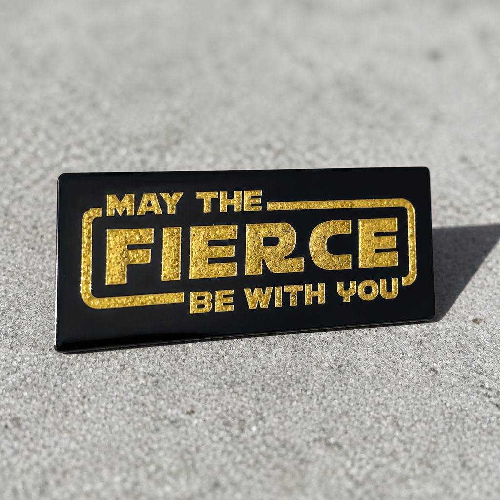 Black enamel pin with glitter gold lettering reading May the Fierce Be With You on a concrete surface