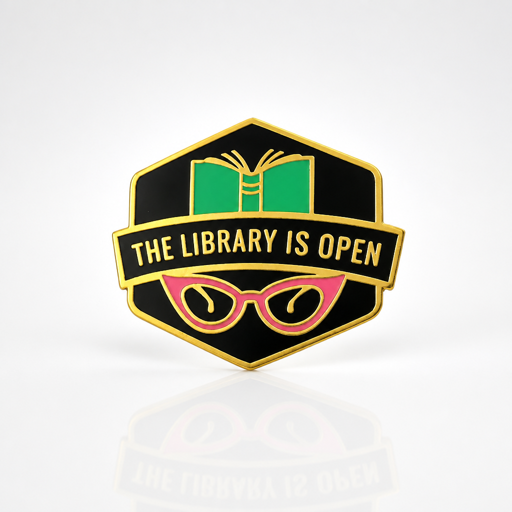 The Library Is Open enamel pin featuring an open book, pink cat-eye glasses, and gold-tone details
