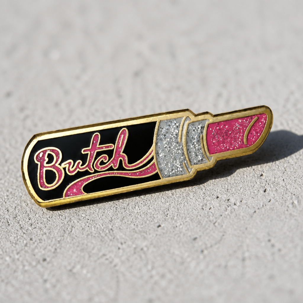 Butch Lipstick enamel pin featuring glitter pink lipstick artwork with gold-tone details on a concrete background