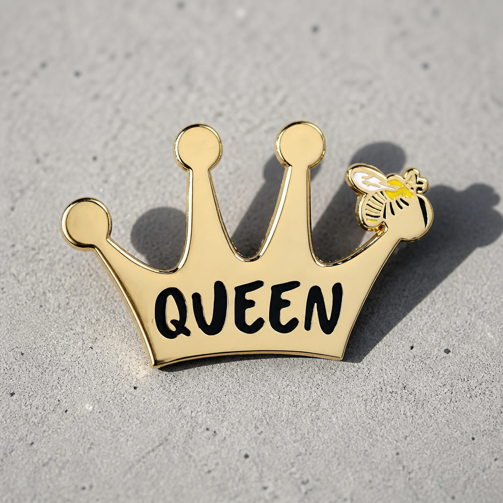 Gold Queen Bee hard enamel pin by Rebellious Pins featuring a crown with a small bee detail and bold Queen lettering displayed on a concrete surface