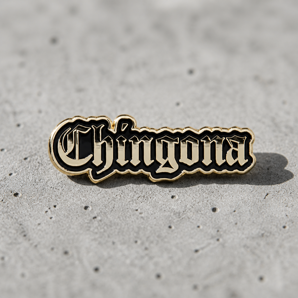 Black and silver Chingona script enamel pin with gothic-style lettering on a concrete surface