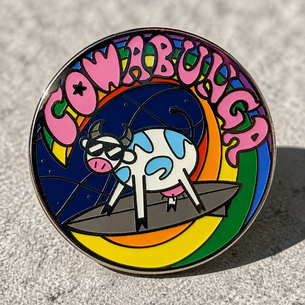 Cowabunga Pride enamel pin with sunglasses-wearing cow surfing on a rainbow wave displayed on a concrete surface