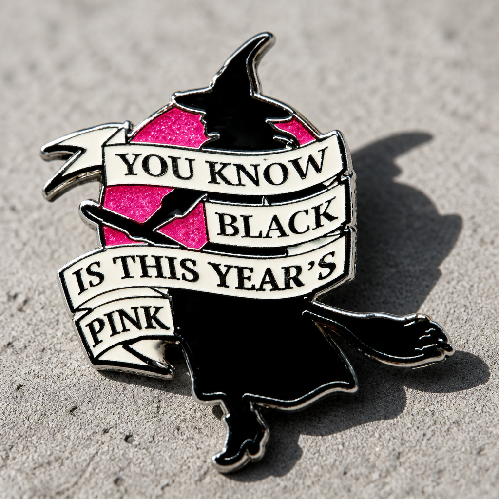Glitter enamel pin featuring a witch silhouette flying across a pink moon with the phrase Black Is This Year’s Pink on a concrete surface