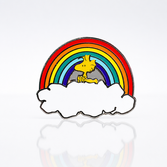 Official Peanuts Woodstock enamel pin featuring Woodstock standing beneath a colorful rainbow resting on white clouds.