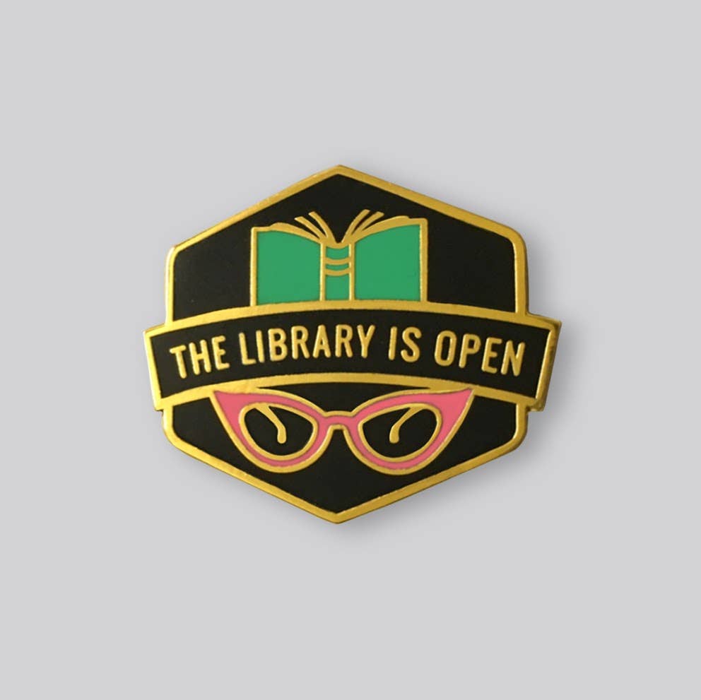 The Library Is Open enamel pin featuring an open book, pink cat-eye glasses, and gold-tone details