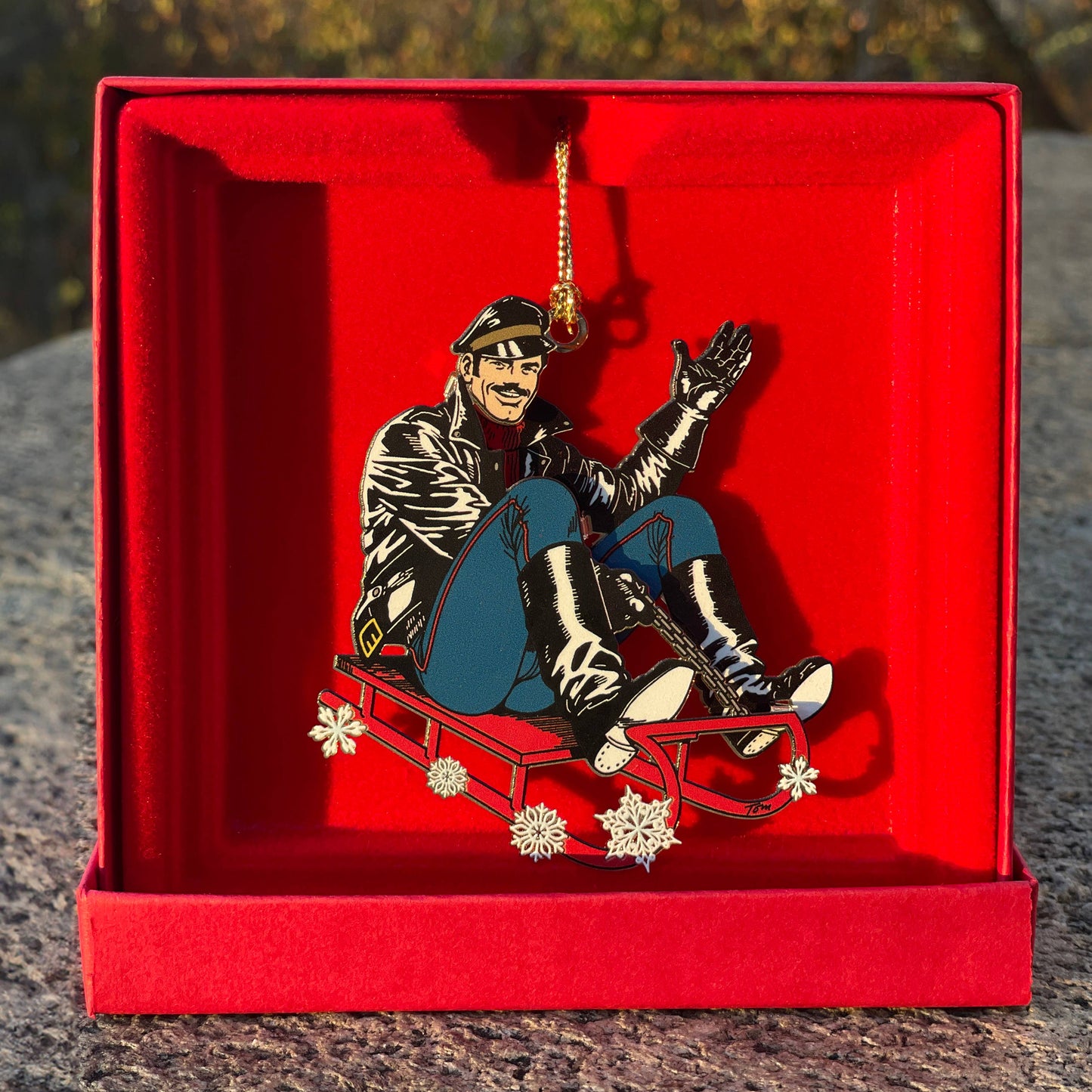 Limited edition Tom of Finland leather sled metal ornament featuring a leather-clad character riding a red sled, displayed in an embossed red gift box.