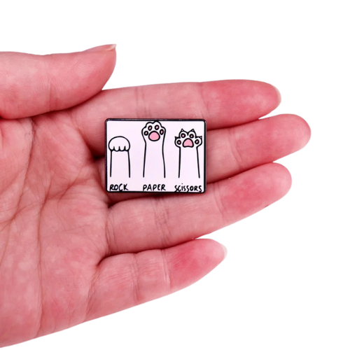 Pin with 'Rock Paper Scissors' design held in a hand on a white background