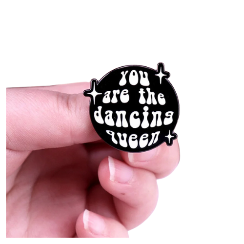 Retro Dancing Queen enamel pin with bold disco-inspired lettering and sparkles held between fingers on a white background