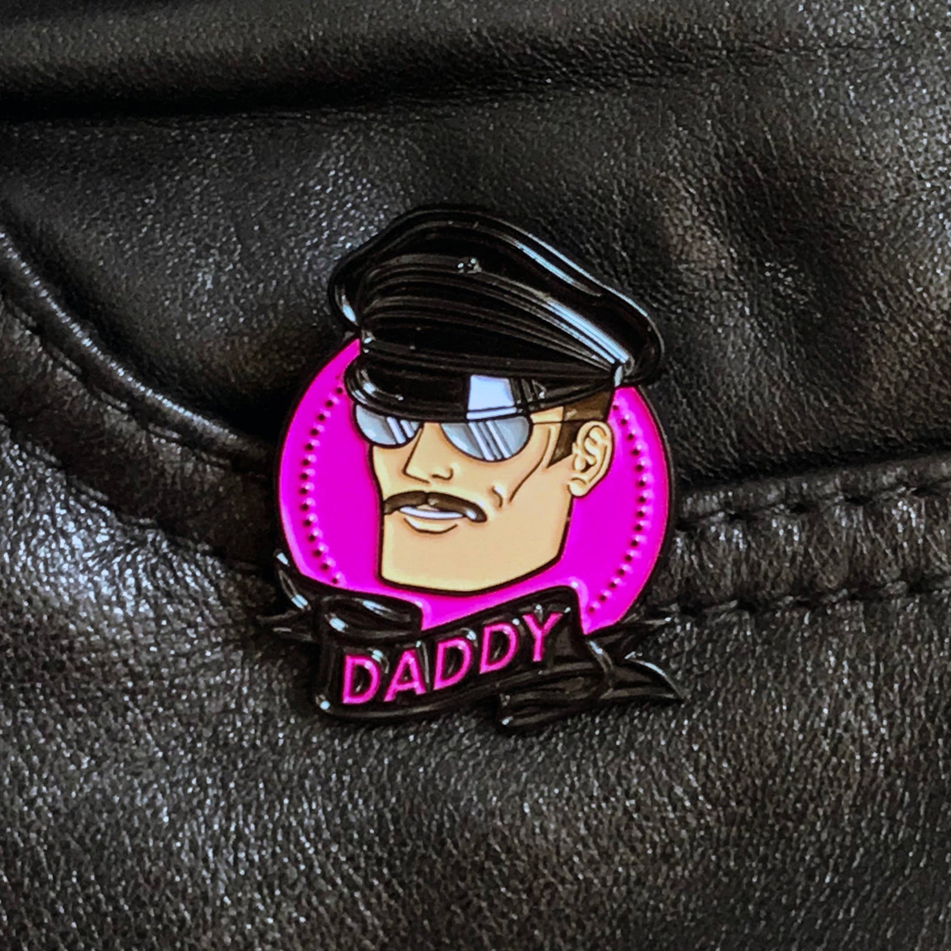 Hard enamel Daddy pin featuring a leather-inspired character wearing sunglasses against a bright pink background.