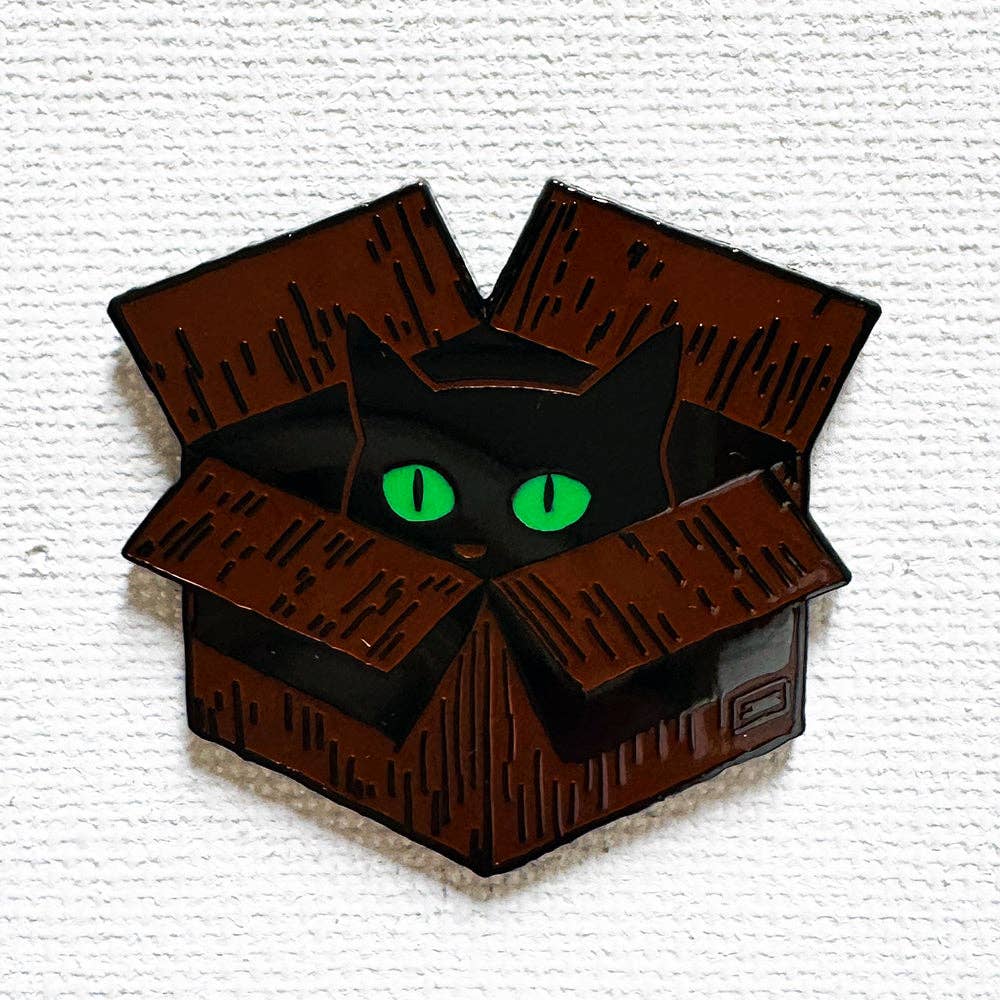 Black cat enamel pin with bright green eyes peeking out from a cardboard box