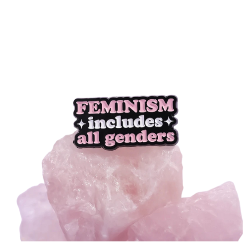 Pink and black enamel pin reading “Feminism Includes All Genders”