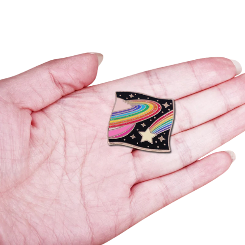 Hand holding a Rainbow Galaxy enamel pin with glittery cosmic rainbow planet and star design