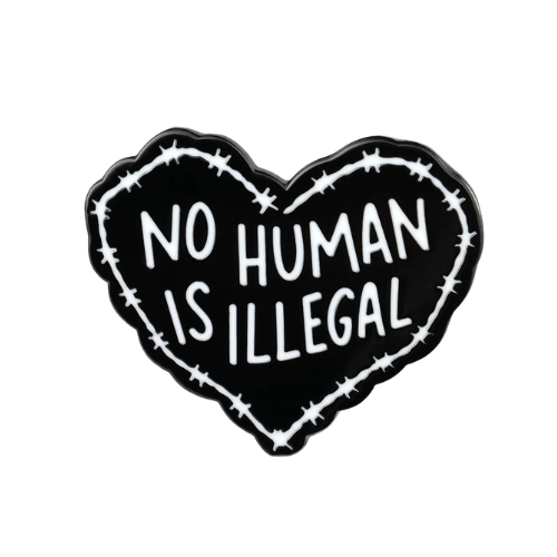 Black heart-shaped enamel pin with the phrase “No Human Is Illegal” in white lettering